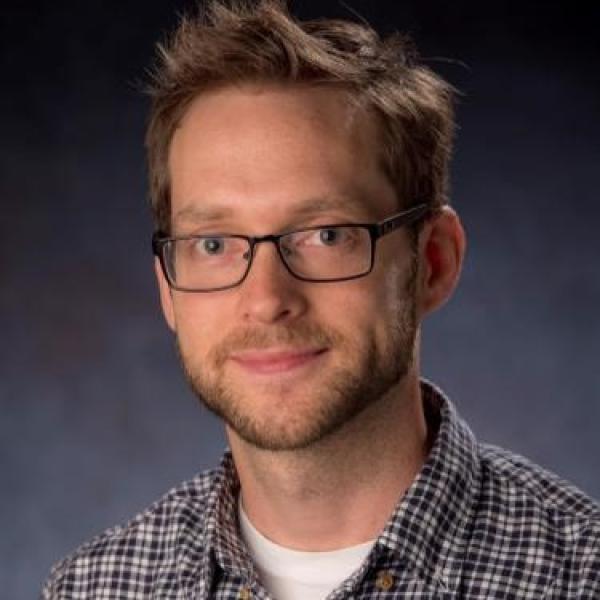 White man with brown hair, dark glasses and facial hair wearing a patterned button-up shirt