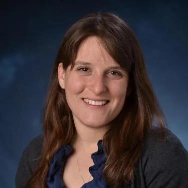 White woman, smiling, with long grown hair wearing a navy ruffled shirt and sweater