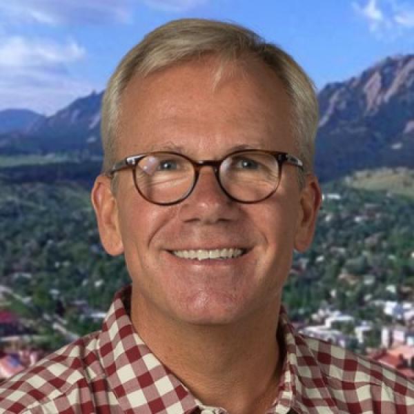 White man with brown hair, round glasses, and red/white plaid collared shirt in front of background photo of Boulder, Colorado