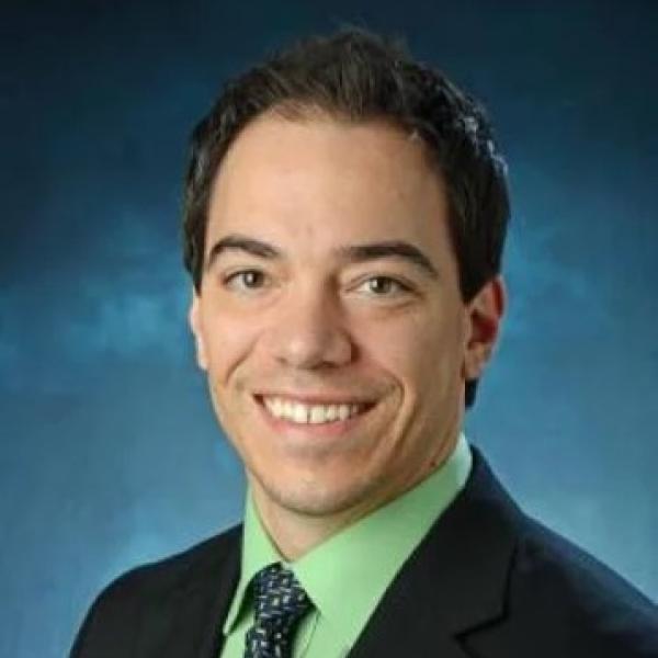 White man wearing a suit jacket and bright green shirt in front of blue background