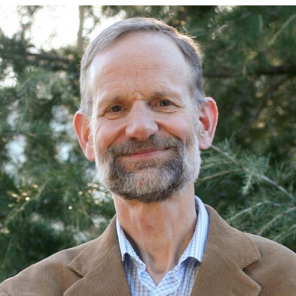 White man with receding hair and full facial hair standing in front of pine tree wearing brown blazer and a blue and button up shirt