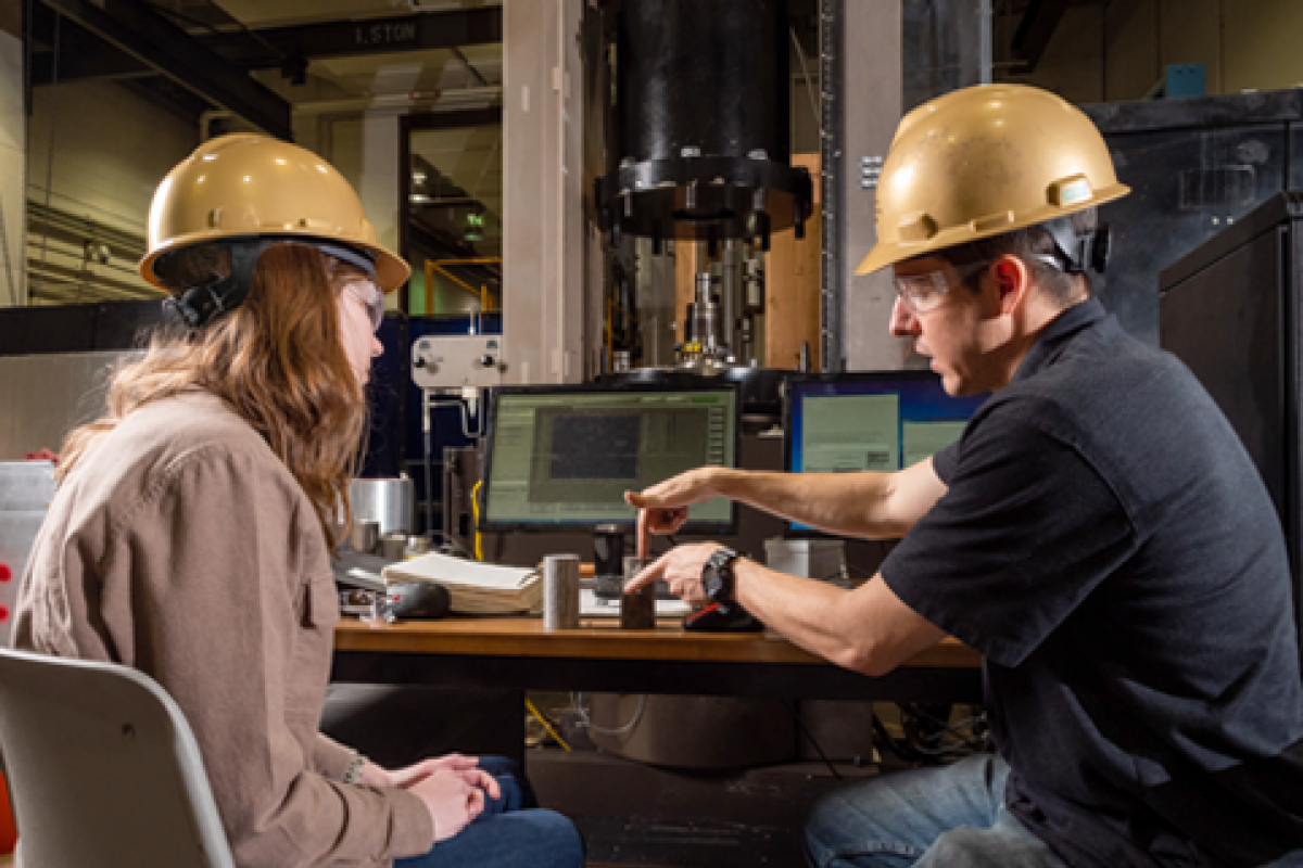 Two students with hard hats on discussing in an engineering lab