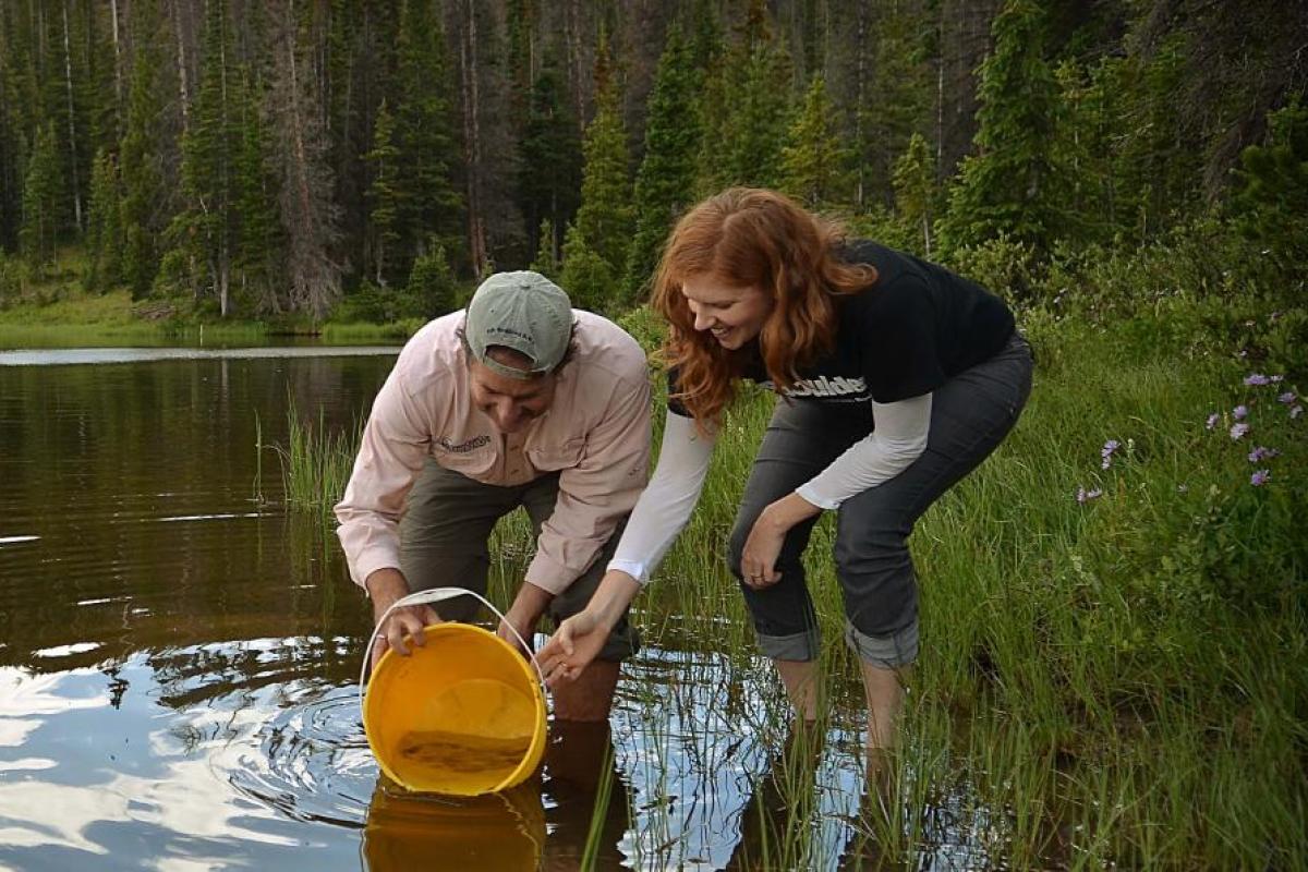 Two smiling people hold a bucket in a large lake