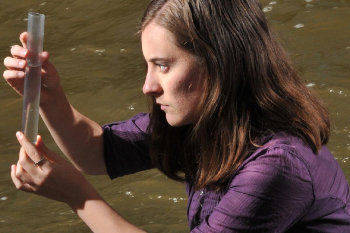 long haired person in purple shirt looks intently at a test tube by a river