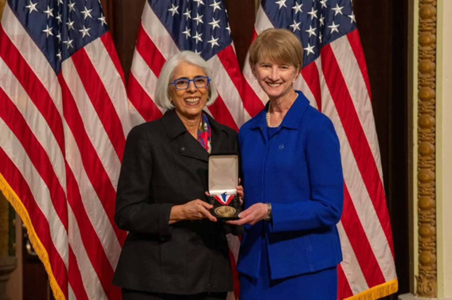 Two woman pose with a medal in front of American flags