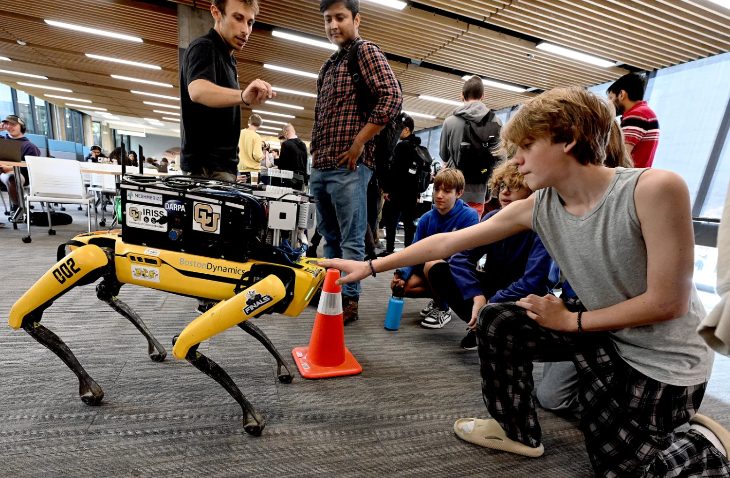Male middle school student, shown kneeling, reaching out to pet a robotic dog.