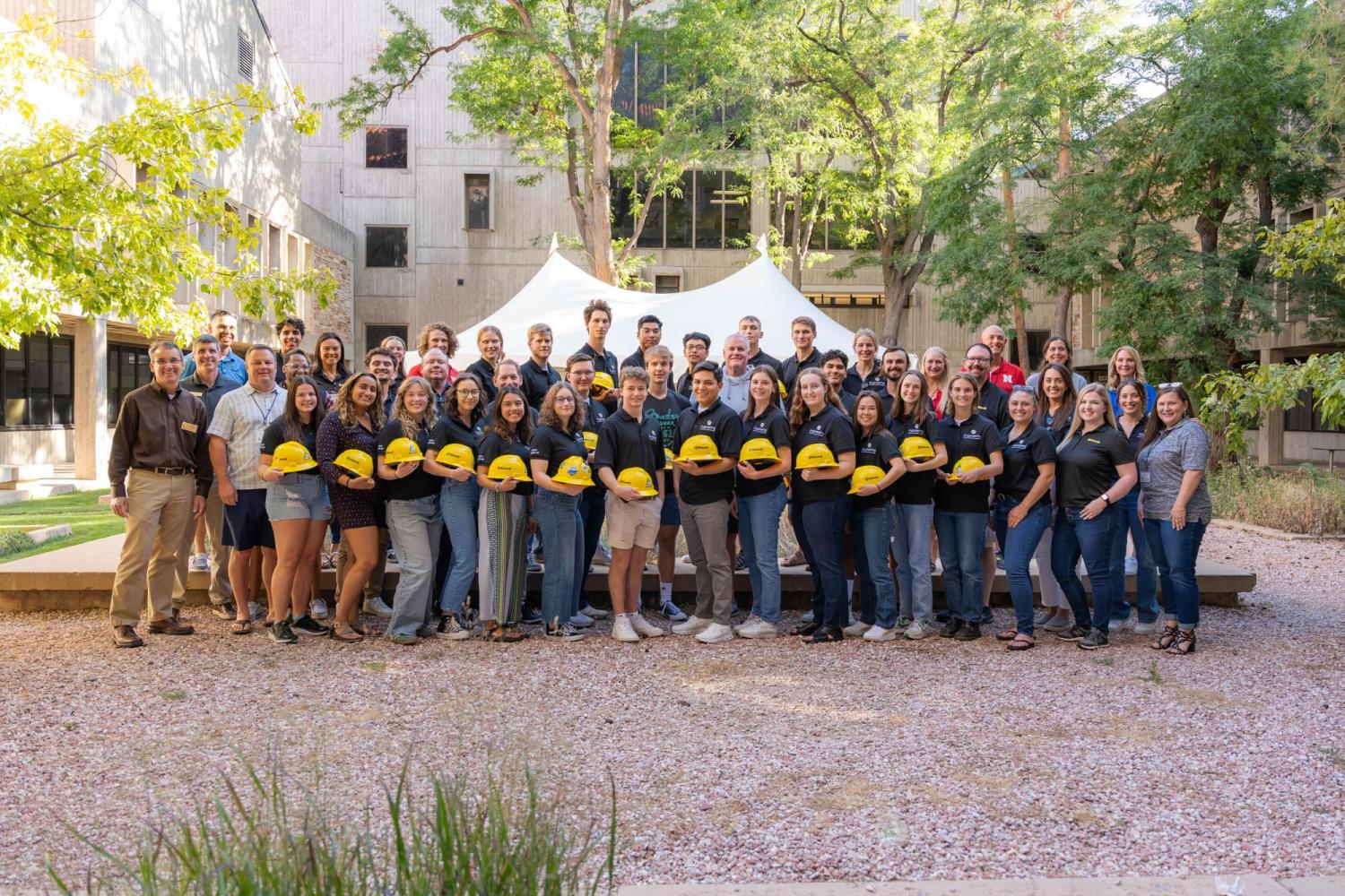 Large group of CU Engineering Kiewit scholars standing in front of engineering building holding a yellow hard hat.
