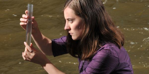 long haired person in purple shirt looks intently at a test tube by a river