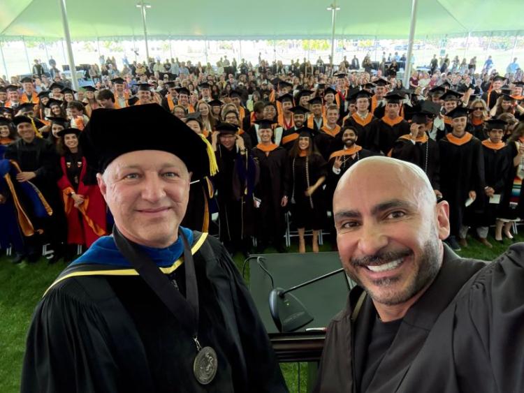 Two individuals in academic regalia smiling at the camera, with a crowd of graduates in caps and gowns seated in the background under a large tent.