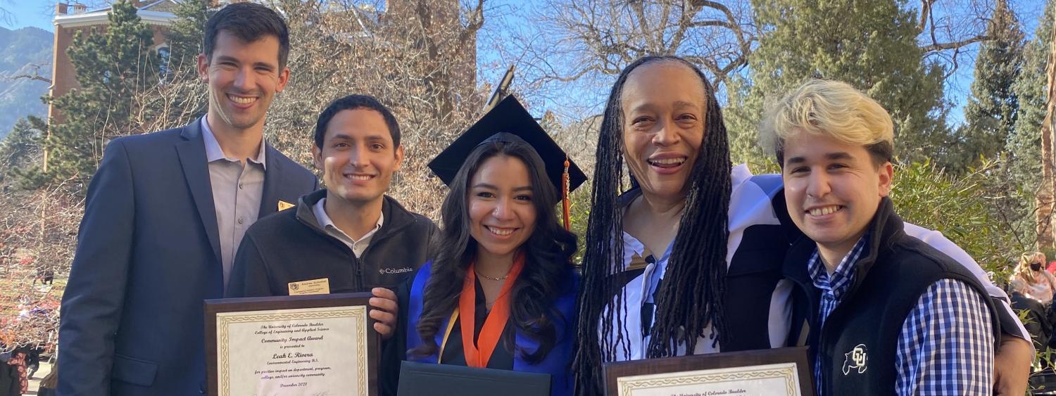 Leah stand in full graduation regalia in the center of four BOLD Staff members helping her hold her two awards under a bright blue sky.