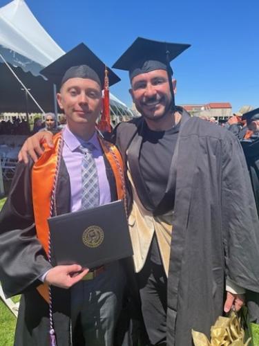 Two individuals in graduation regalia, one holding a diploma, smiling at the camera.
