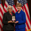 Two woman pose with a medal in front of American flags