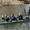 Five students in wet suits in their concrete canoe on the water. The person in the back raised his paddle up with two hands.