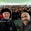Two individuals in academic regalia smiling at the camera, with a crowd of graduates in caps and gowns seated in the background under a large tent.