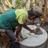 A man hunched down over a concrete pit for an alternating dual-pit latrine holding a tool