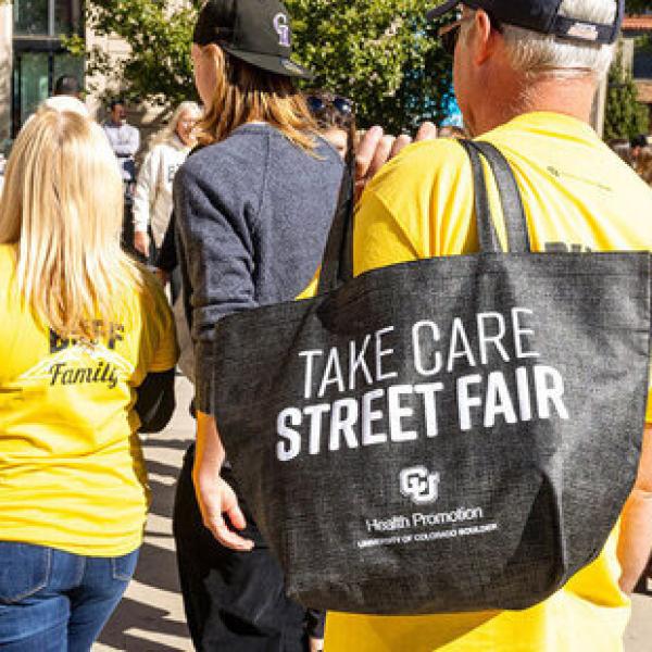 people walk with 'Take Care Street Fair' tote bags