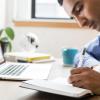 person writing in notebook and working on computer