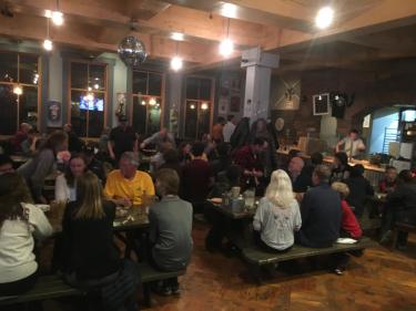 Lots of people sit at indoor picnic tables with pizza and drinks.