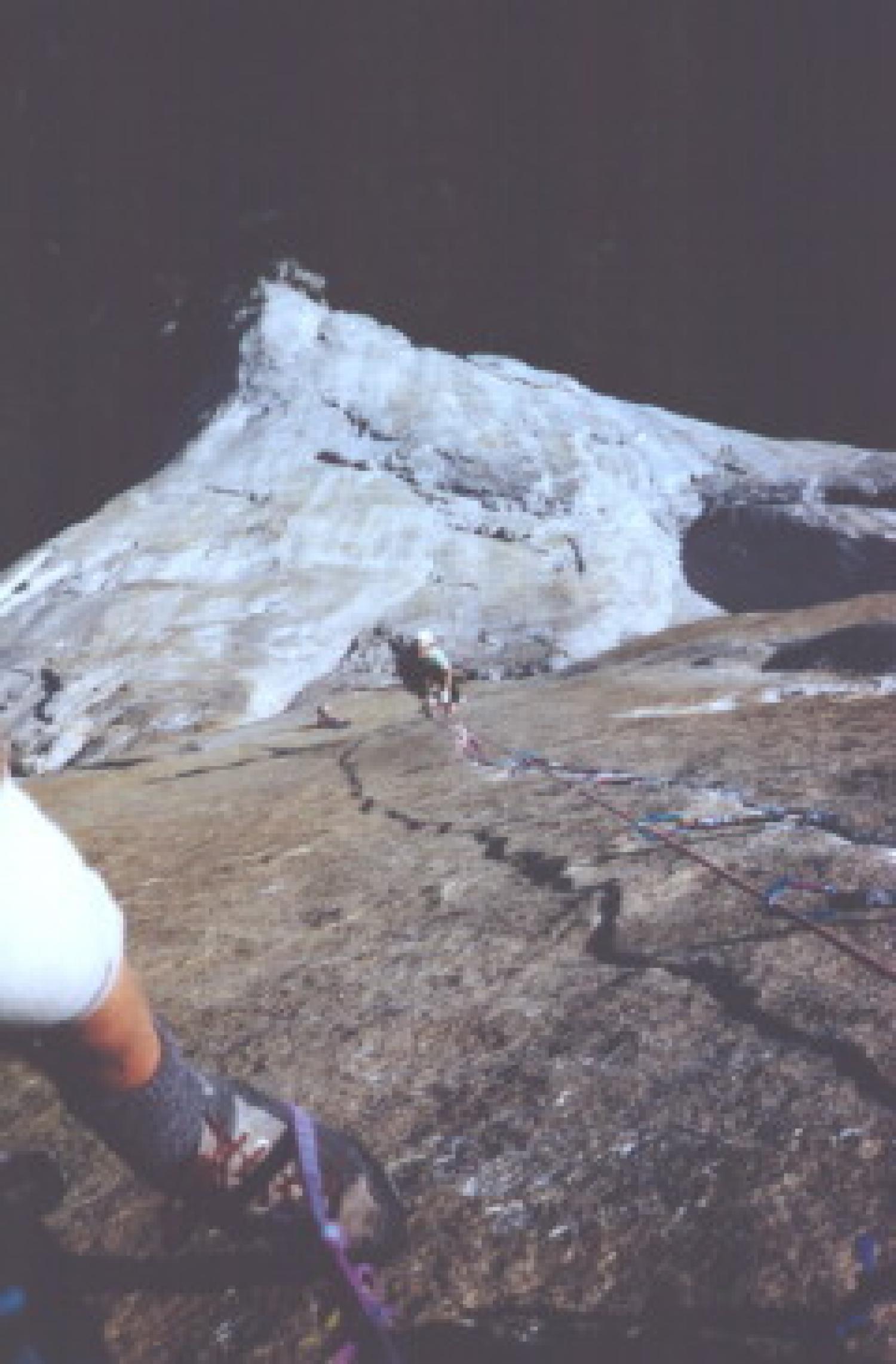 View from above looking down at someone rockclimbing up. The photographers foot is visible against the rock face.