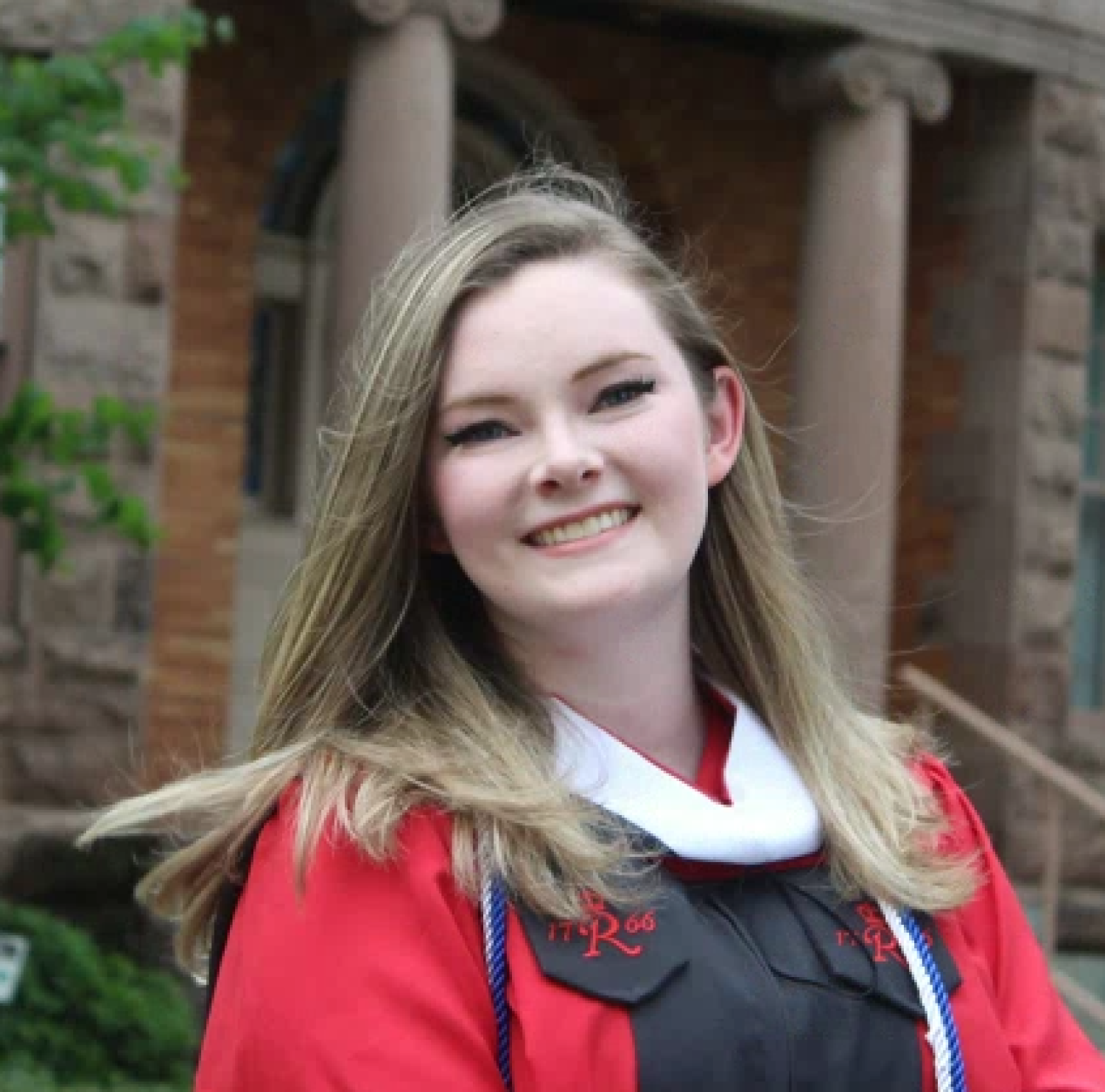 Georgia in a red graduation gown smiling in front of a brick building