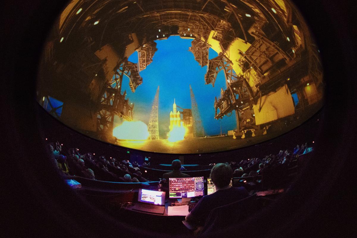 Interior of dome showing a rocket launch on the screen, operator's console in view too