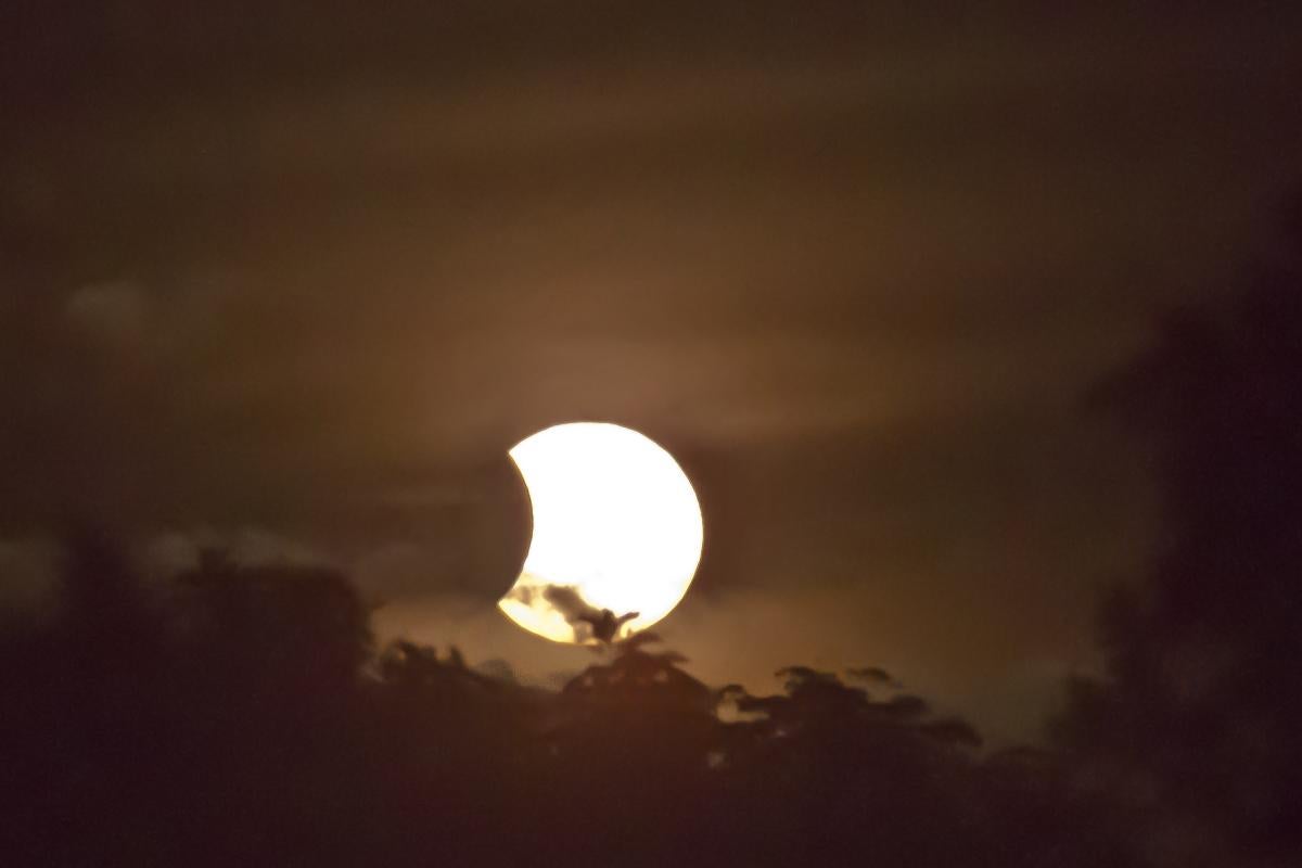 Beginning of a total solar eclipse with moon just moving over corner of the sun clouds and trees on horizon