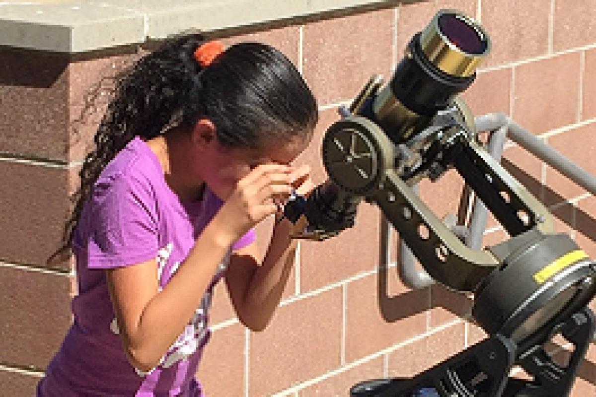 Girl standing by a brick wall looking through a solar telescope