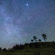 Photo of the night sky with grassy hill and trees and a meteor in the sky