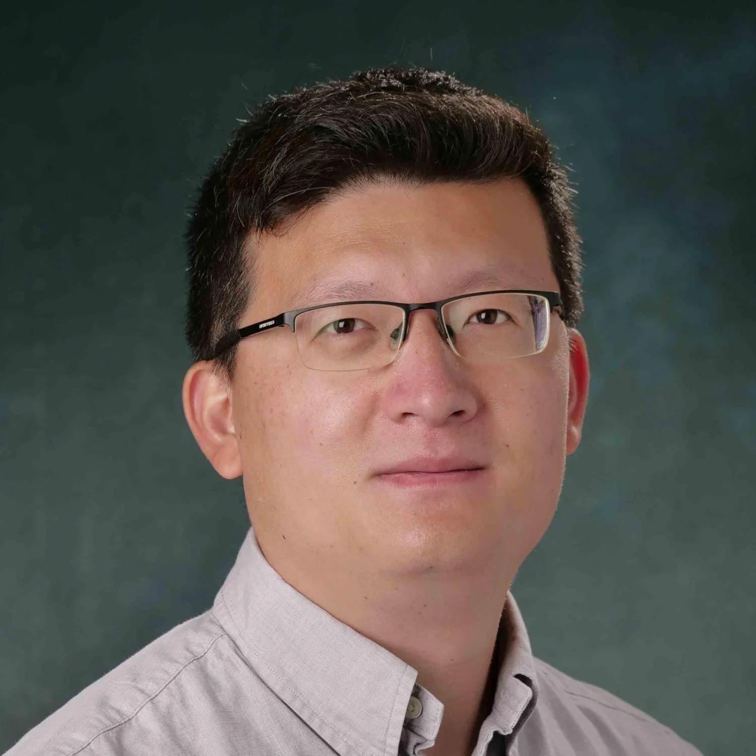 Yida Zhang wearing glasses and a button-down shirt in front of a green background.