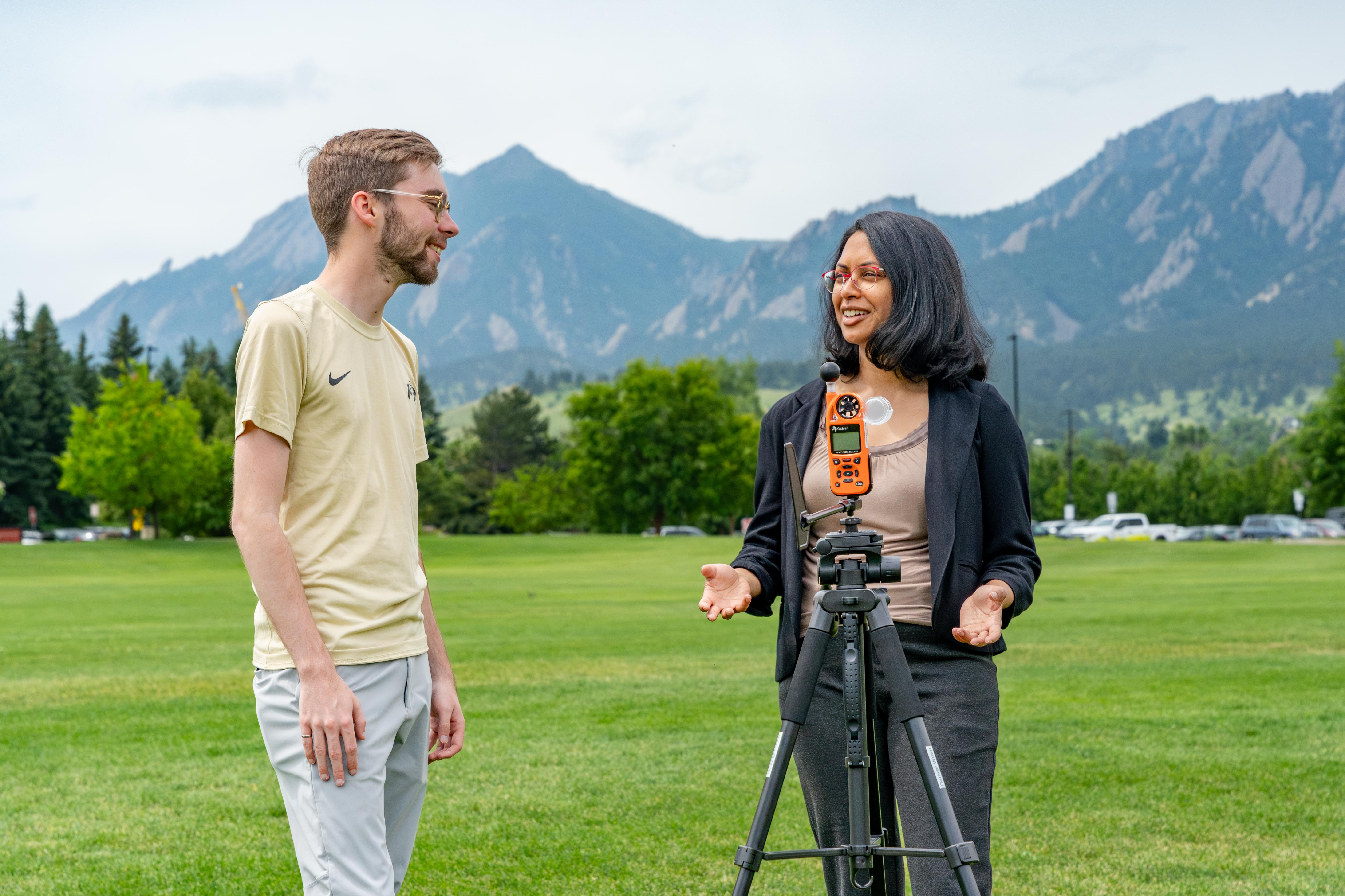 Aditi Bhaskar and student interact by a hydrology instrument in front of a large expanse of lawn with the Flatirons in the background.