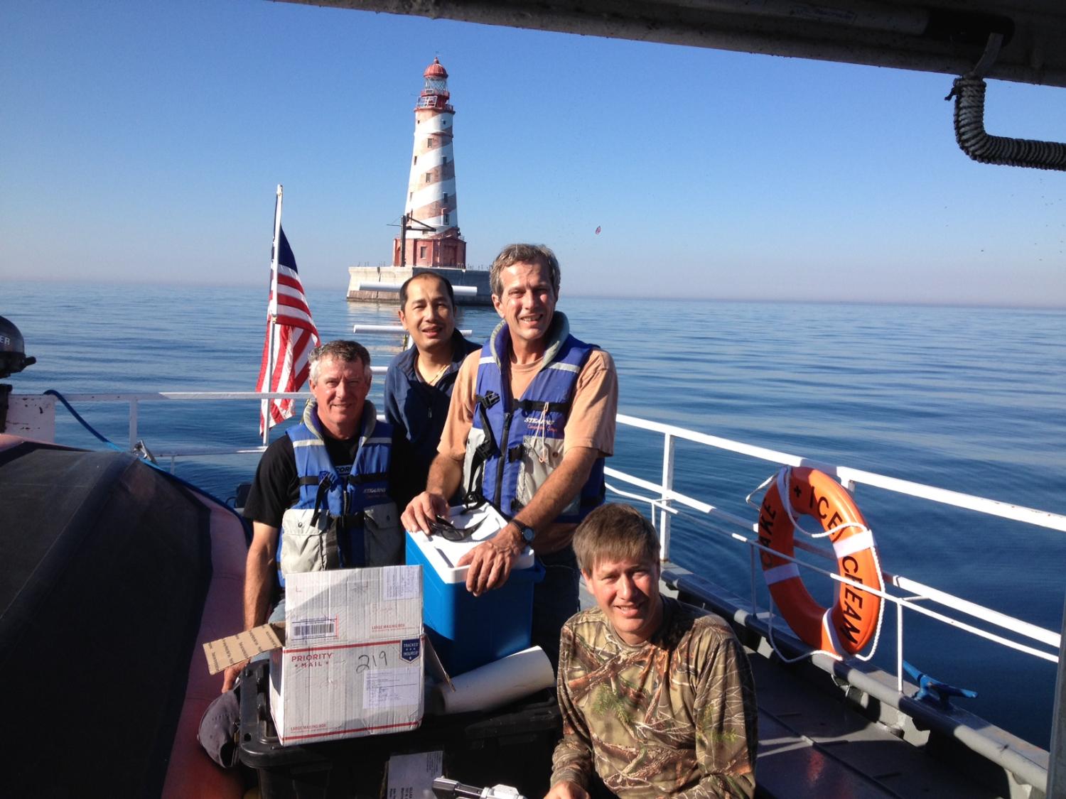 4 men on deck of boat in front of a lighthouse posing for photo