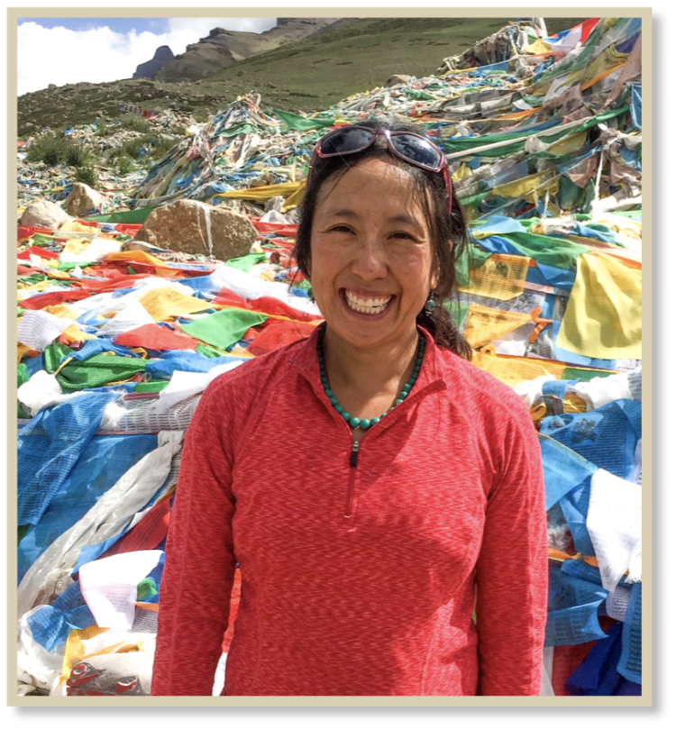 Emily Yeh in foreground with Tibetan landscape and prayer flags on the ground behind her.