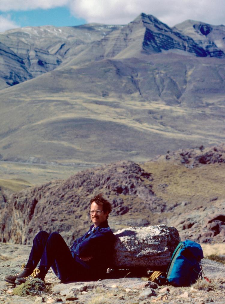 Man sitting on ground, leaning against a rock with mountain landscape in the background