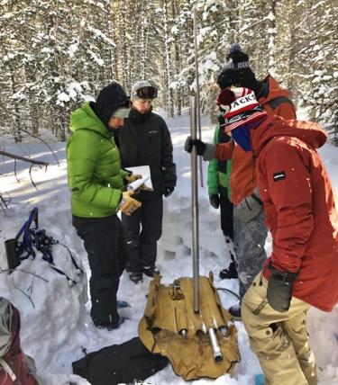 4 people in winter coats & hats standing in the snow, looking down at research equipment