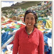 Emily Yeh in foreground with Tibetan landscape and prayer flags on the ground behind her.