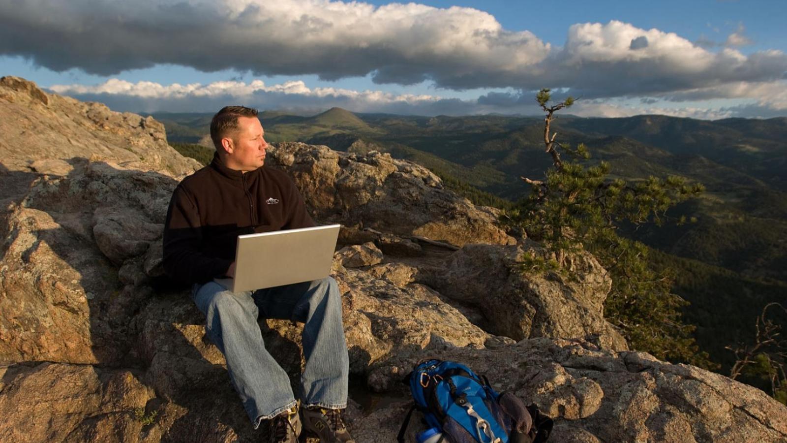 Man with laptop on a rocky overlook in the mountains