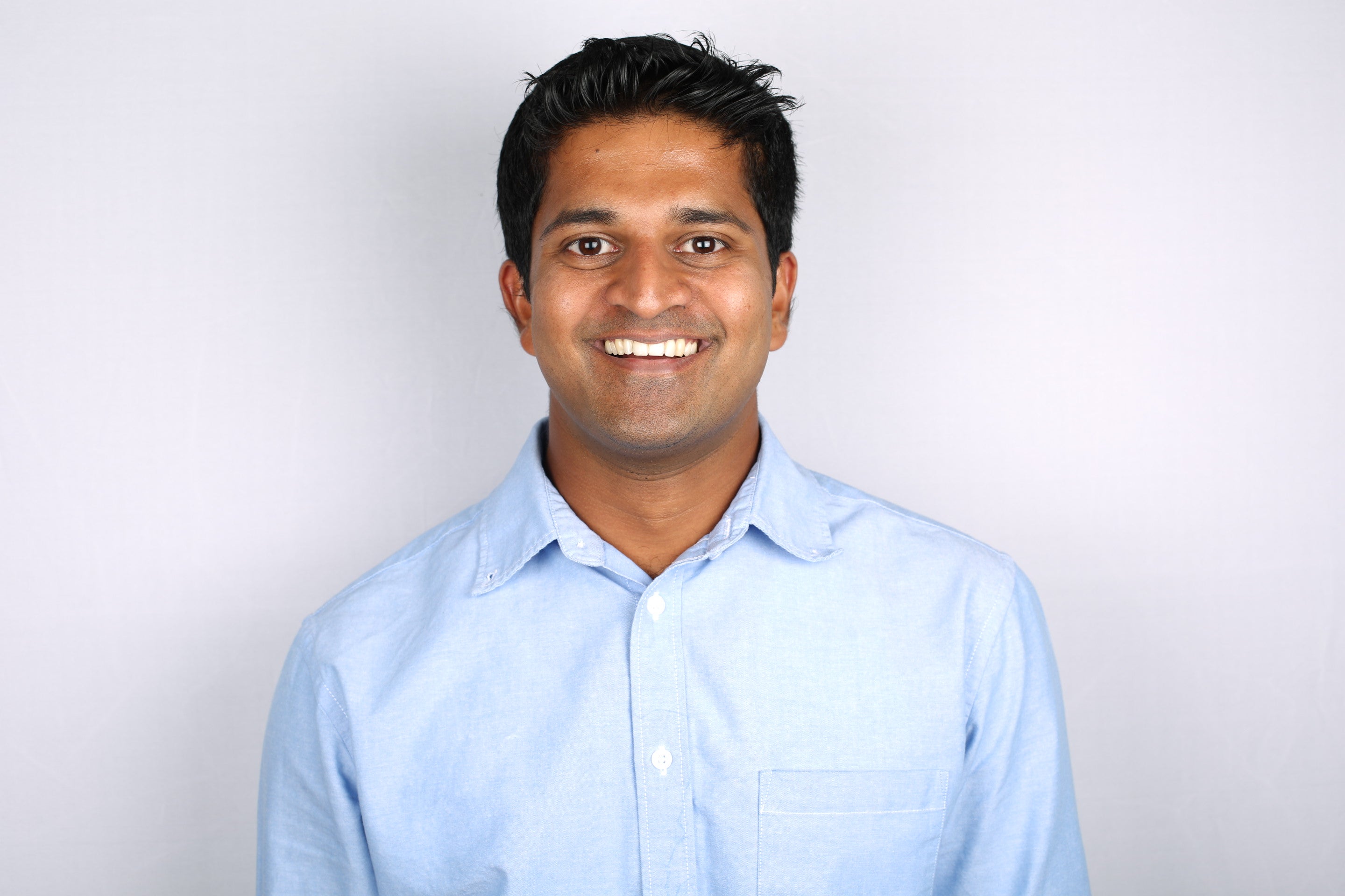 A headshot of a smiling man wearing a light blue shirt and black hair
