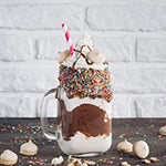 Mason jar filled with ice cream, syrup and sprinkles on a table with cookie crumbles. 