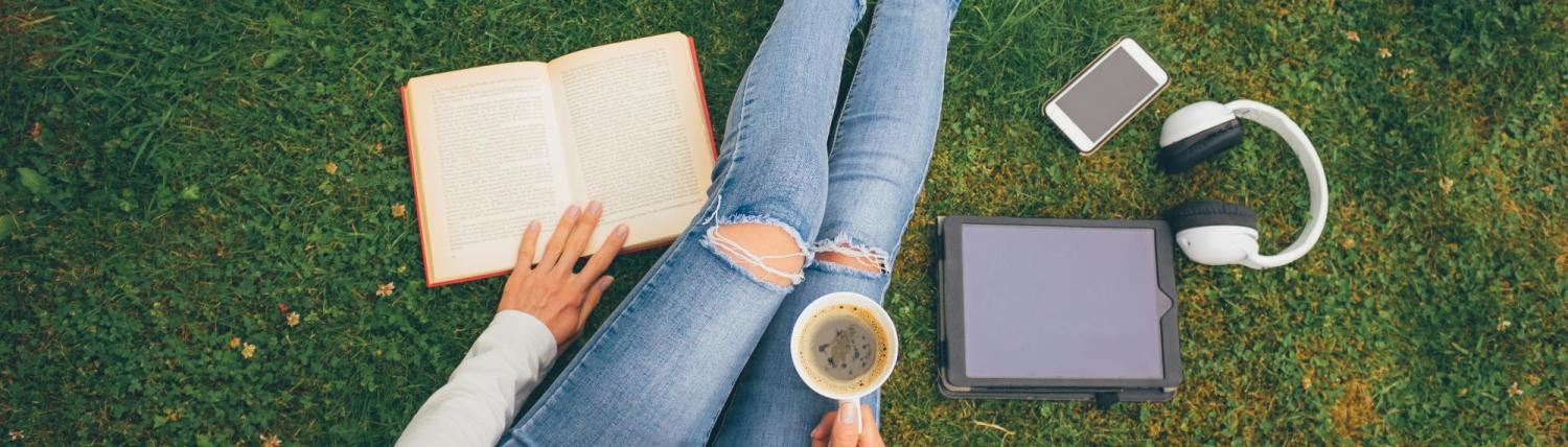 Person laying in the grass with headphones, a laptop and coffee