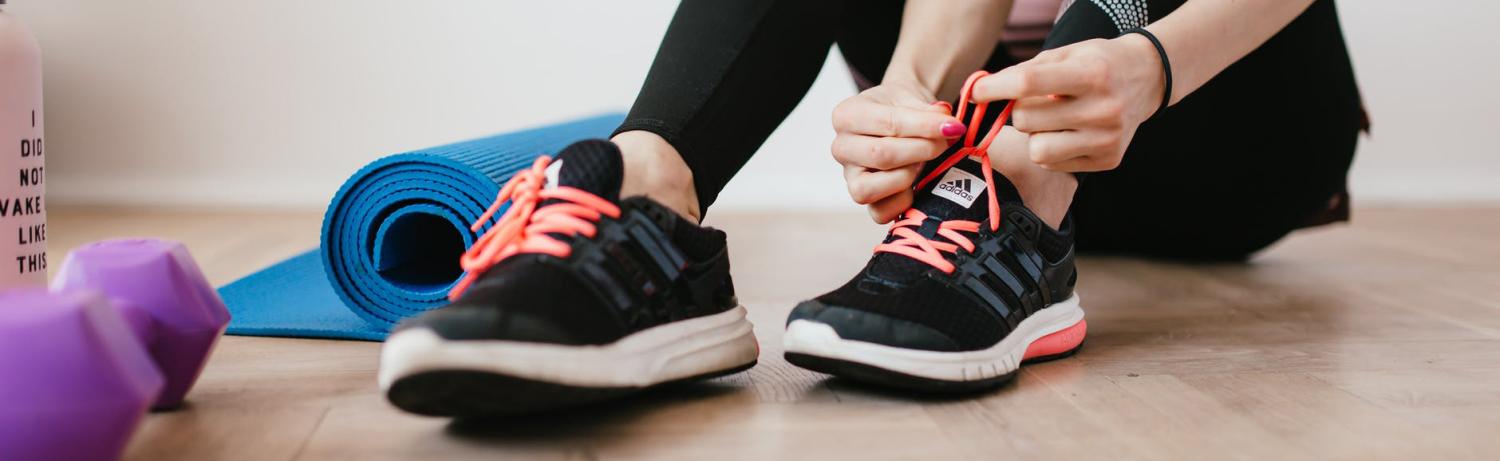 Person lacing up black tennis shoes next to a yoga mat, dumbbells and a water bottle on the floor.