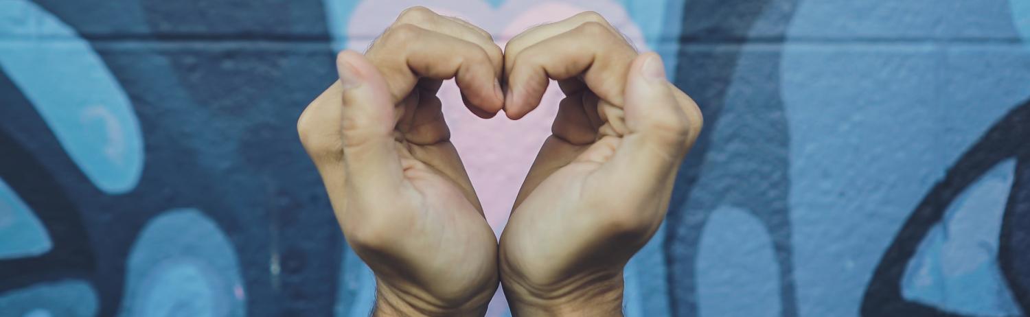 Person holding up their hands in a shape of a heart in front of a colorful outdoor mural.