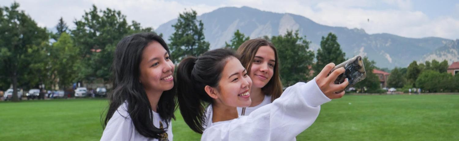 Three students taking a selfie together in front of the flatirons by farrand field.