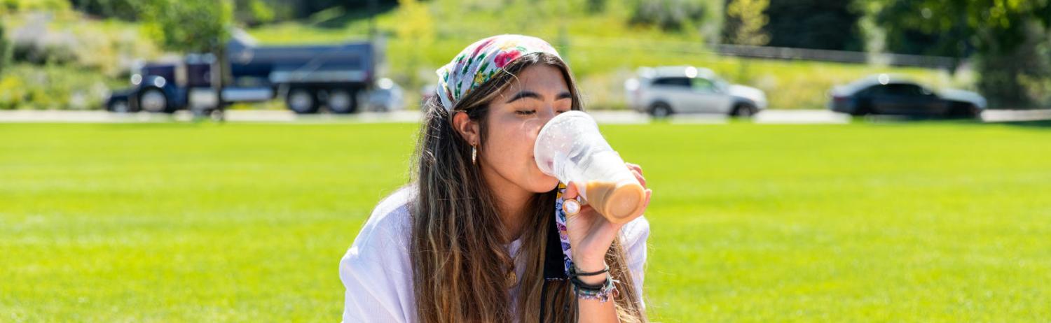 Photo of a student sipping on a coffee and enjoying a sunny day on the Business Field.
