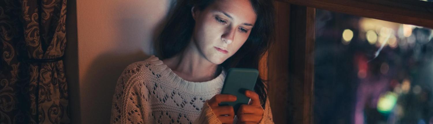 Photo of a girl sitting in a dim lit room looking at her phone with concern.