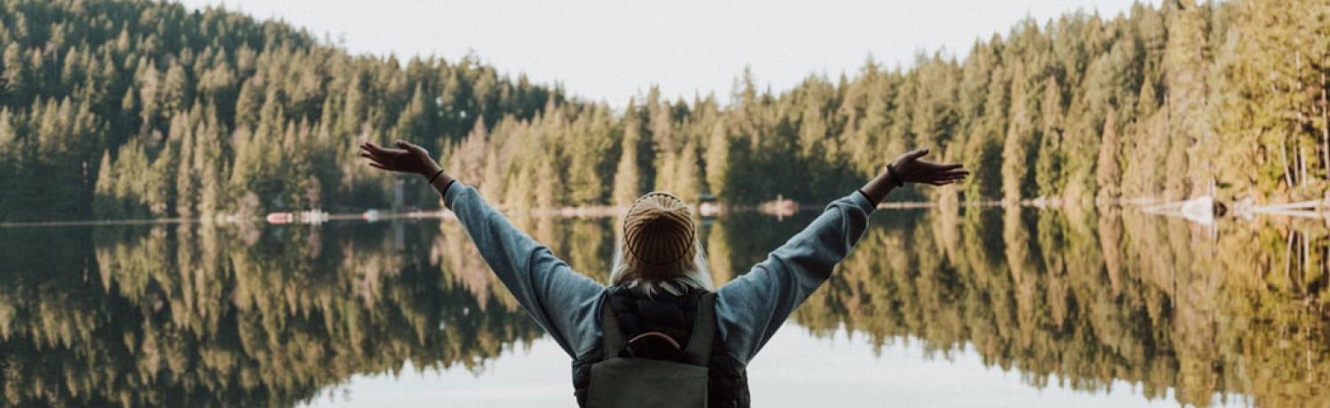 Girl wearing a beanie holding her arms up with joy in front of a forested lake.