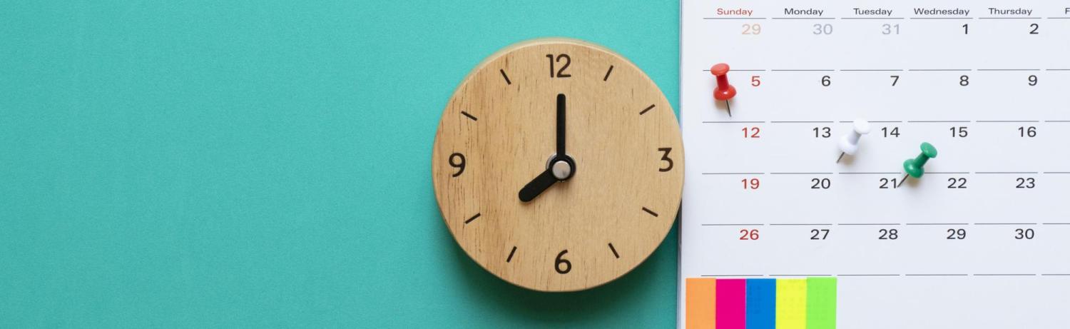 Wooden clock sitting next to a spiral calendar with colorful tacs.