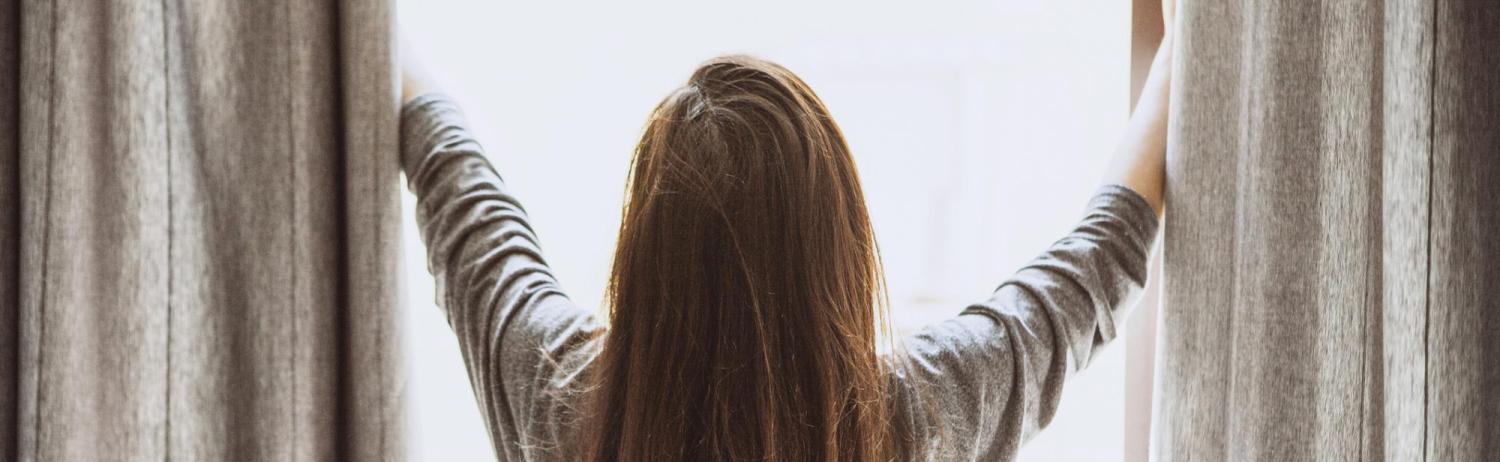 Girl standing in front of her window as she pulls back the blinds and lets the sunlight in.