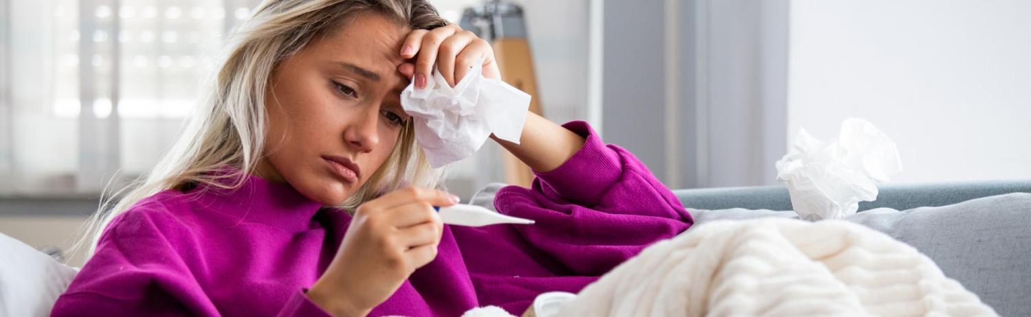 An ill looking girl holding a fistful of tissues to her forehead as she peers down at a thermometer.