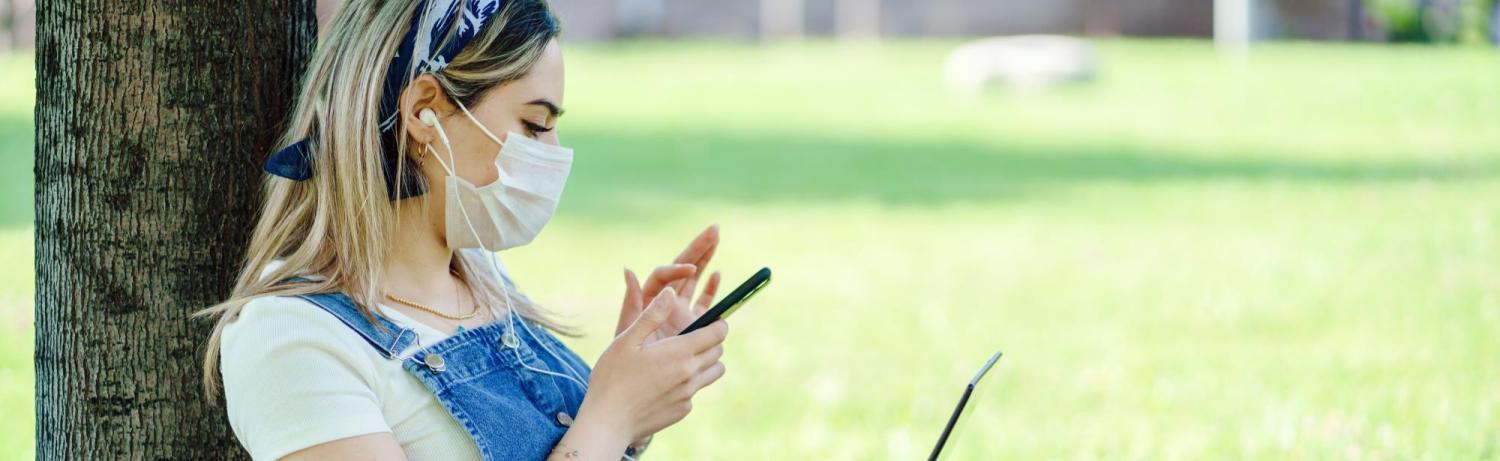 Female student in mask checking her phone outside while sitting under a tree.