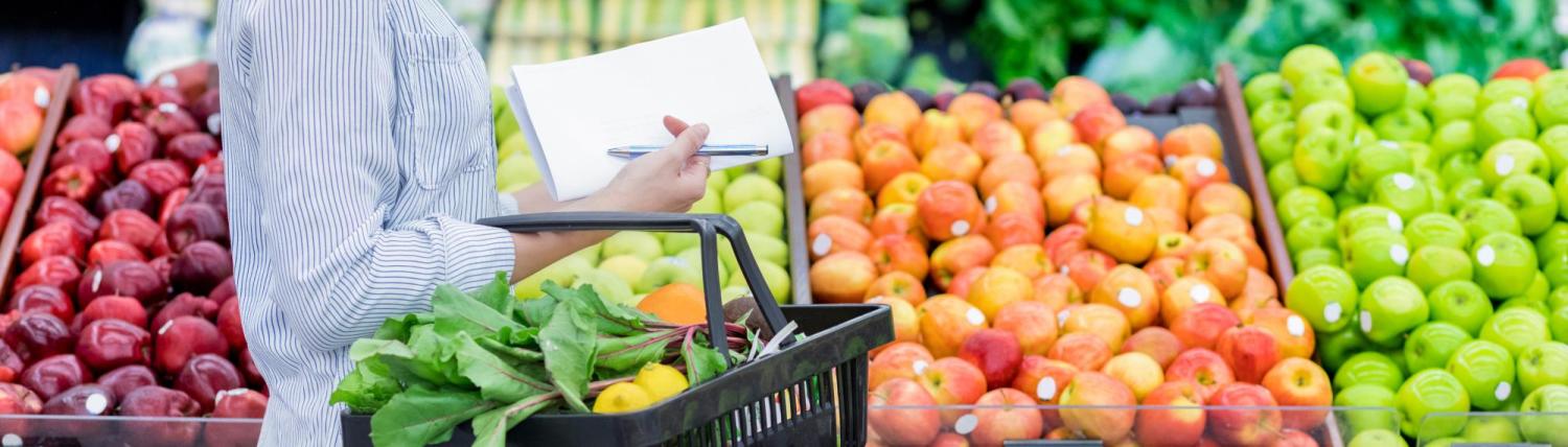 Woman holding a list and grocery basket while standing in front of a produce bin.